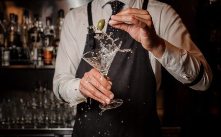 Bartender holding a splashing fresh and strong summer martini cocktail and a green olive against the bar