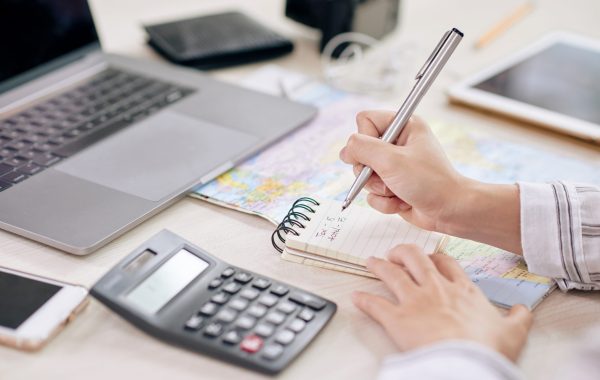 Crop shot of woman taking notes in notepad sitting at desk with laptop and calculating machine